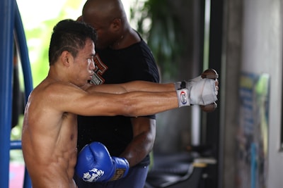 A person engaging in boxing training, wearing boxing gloves and hand wraps, with a coach or trainer standing nearby for assistance. The environment appears to be a gym with equipment visible, including a boxing ring.