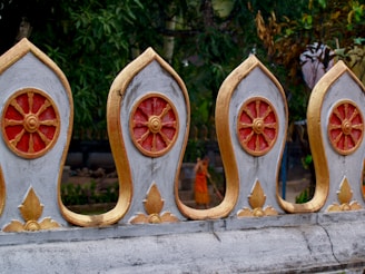 A beautifully crafted ornate fence with circular, wheel-like motifs painted in gold and red. The background reveals lush green foliage and a person wearing orange robes standing behind the fence.