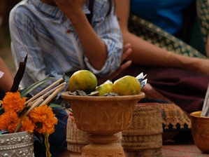A close-up of a ceremonial setup with a wooden pedestal holding citrus fruits and pieces of paper. Marigold flowers and incense sticks are placed beside in a silver container. Behind, people are seated, wearing traditional woven fabric.