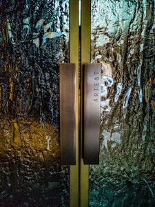 A pair of vertical metallic handles are fixed onto textured glass doors with a wavy pattern. The glass surface has reflective qualities with shades of teal and amber. The word 'ARTEST' is engraved on one of the handles.