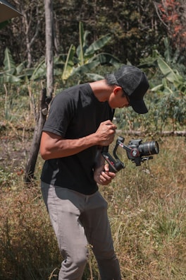 A person wearing a black cap, black t-shirt, and gray pants is operating a camera mounted on a stabilizer. The setting is an outdoor grassy area with green foliage in the background, including banana plants.