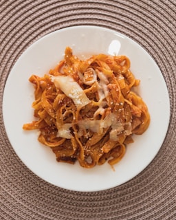 A plate of pasta with a rich tomato-based sauce is topped with melted cheese and garnished with grated Parmesan. The pasta sits on a white plate, which is placed on a textured, circular placemat.