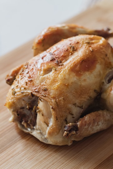 A roasted chicken with golden brown, crispy skin, seasoned with herbs, and placed on a wooden cutting board.