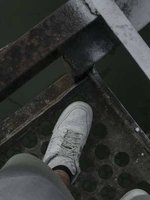 A white sneaker is placed on a perforated metal surface, with the edge of the surface showing some rust. The color of the metal appears worn and the surface has a grid of circular holes. The background is a dark green color, possibly water.
