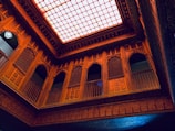 An intricately designed interior with ornate wooden carvings and lattice work on the walls, balconies, and ceiling. The ceiling features a large, illuminated grid pattern that casts a warm light over the space. The architecture includes an array of arches and detailed geometric patterns.