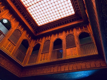 An intricately designed interior with ornate wooden carvings and lattice work on the walls, balconies, and ceiling. The ceiling features a large, illuminated grid pattern that casts a warm light over the space. The architecture includes an array of arches and detailed geometric patterns.