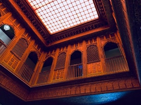 An intricately designed interior with ornate wooden carvings and lattice work on the walls, balconies, and ceiling. The ceiling features a large, illuminated grid pattern that casts a warm light over the space. The architecture includes an array of arches and detailed geometric patterns.