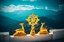 Golden statues of two deer flank a large golden dharma wheel, set against a backdrop of misty blue mountains under a partly cloudy sky. A white terrace provides the foreground.