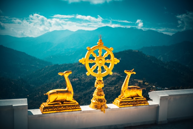 Golden statues of two deer flank a large golden dharma wheel, set against a backdrop of misty blue mountains under a partly cloudy sky. A white terrace provides the foreground.