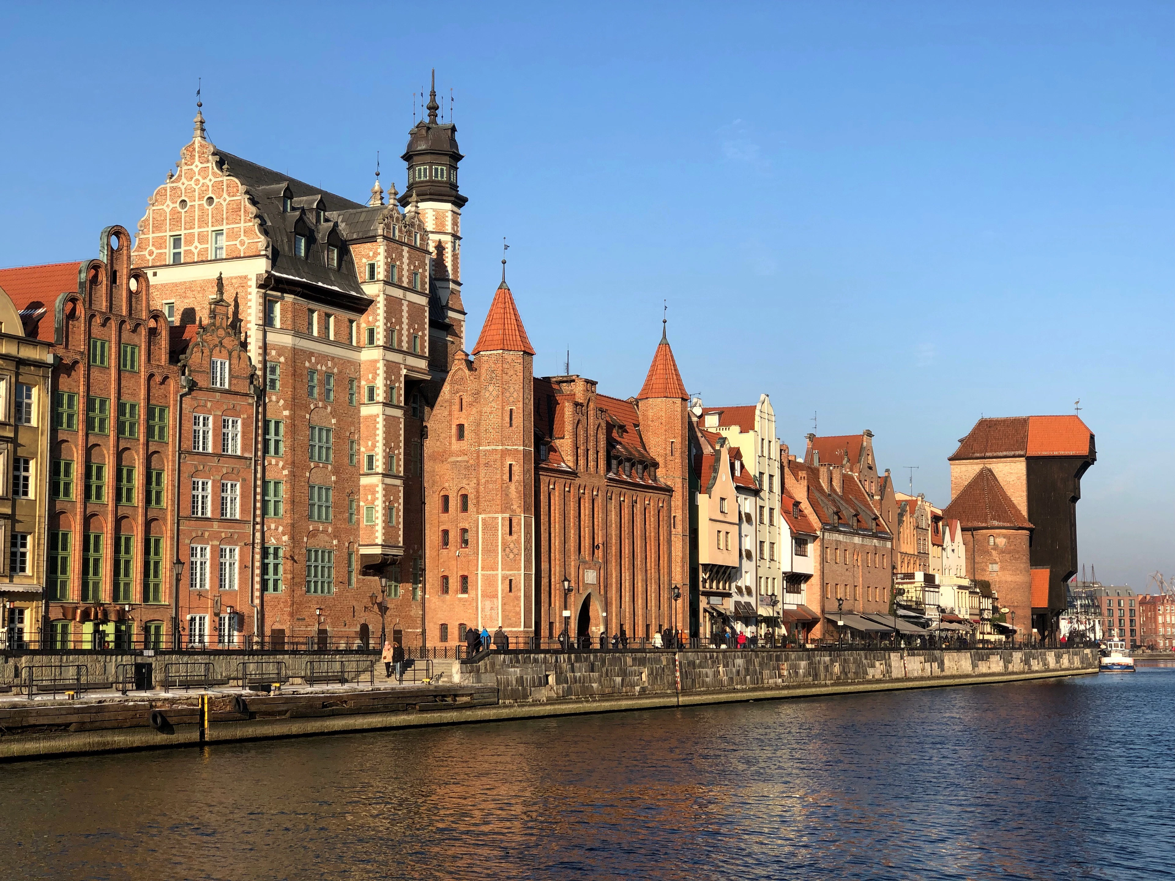 The brick and stone waterfront buildings along the Motlawa river in Gdansk on a clear day, with the dark timber Crane visible downstream