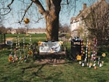 A wooden bench is adorned with a colorful 'Happy Easter' sign and decorated with large, yellow flowers and strings of variously colored handmade ornaments. The bench is situated under a tree with bare branches. In the background, there are quaint stone houses and a grassy field. The entire scene is illuminated by bright daylight, suggesting a cheerful, festive atmosphere.