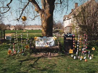 A wooden bench is adorned with a colorful 'Happy Easter' sign and decorated with large, yellow flowers and strings of variously colored handmade ornaments. The bench is situated under a tree with bare branches. In the background, there are quaint stone houses and a grassy field. The entire scene is illuminated by bright daylight, suggesting a cheerful, festive atmosphere.