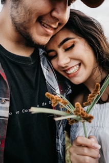 A close-up of two smiling individuals, one holding a small bouquet of flowers. The person on the left is wearing a plaid shirt over a black t-shirt and has facial hair. The person on the right is leaning their head on the other’s shoulder, wearing a white off-the-shoulder top and has long dark hair and eye makeup.