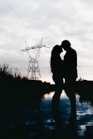 A romantic silhouette of a couple sharing a tender moment near a reflective body of water during twilight. The scene features a large electrical tower in the background and is filled with soft, muted lighting.