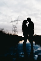 A romantic silhouette of a couple sharing a tender moment near a reflective body of water during twilight. The scene features a large electrical tower in the background and is filled with soft, muted lighting.