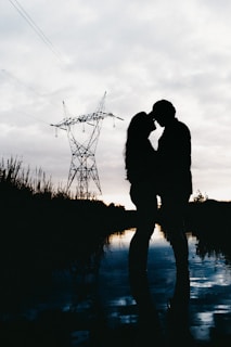 A romantic silhouette of a couple sharing a tender moment near a reflective body of water during twilight. The scene features a large electrical tower in the background and is filled with soft, muted lighting.