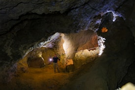 A person holding a flashlight explores a dimly lit cavern with rugged rock formations. The cave features various natural textures, with light casting shadows on the walls and floor.