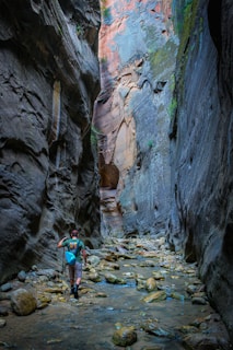 A person hiking through a narrow canyon with towering rock walls. The ground is covered with water and large stones, and the hiker is using trekking poles and carrying a small backpack. The walls have various shades of grey and brown, and there are patches of vegetation growing on them.