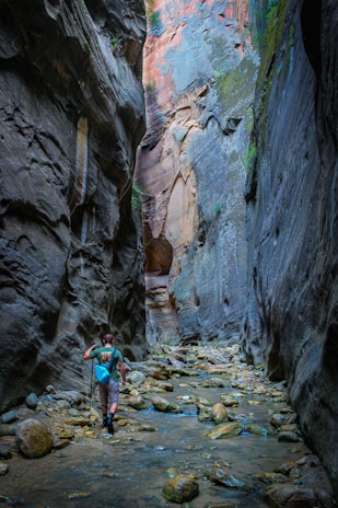 A person hiking through a narrow canyon with towering rock walls. The ground is covered with water and large stones, and the hiker is using trekking poles and carrying a small backpack. The walls have various shades of grey and brown, and there are patches of vegetation growing on them.