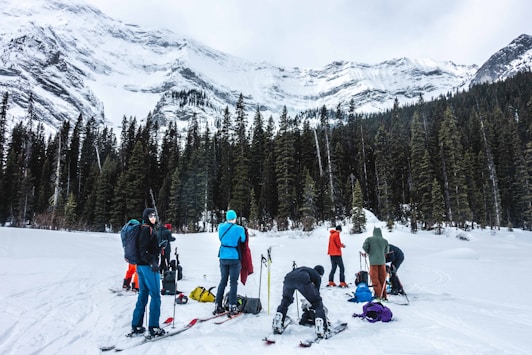 A group of people is preparing for skiing surrounded by snow-covered mountains and tall evergreen trees. They are equipped with skis and backpacks, and the sky is overcast, suggesting cold weather conditions.