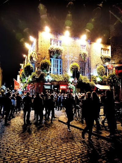 A vibrant street scene at night featuring a lively crowd gathered outside a brightly lit pub adorned with hanging plants. The cobblestone street reflects the lights, and the building is illuminated with a warm glow, creating an inviting atmosphere.