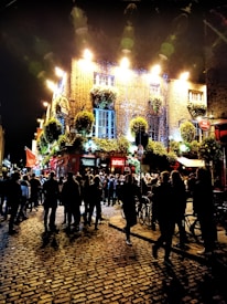 A vibrant street scene at night featuring a lively crowd gathered outside a brightly lit pub adorned with hanging plants. The cobblestone street reflects the lights, and the building is illuminated with a warm glow, creating an inviting atmosphere.
