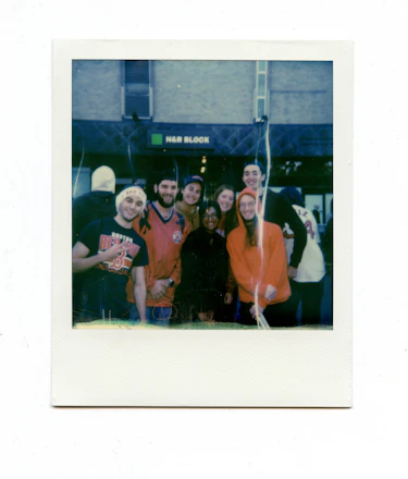 A group of six people, some wearing sports jerseys and hats, are standing in front of an H&R Block storefront. They appear to be happy and possibly celebrating an event together.