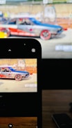 A colorful race car is displayed on a screen and simultaneously captured on a smartphone. The car features vibrant red and blue colors with decals. It is parked in what seems to be a race track environment with barriers and indistinct background structures.