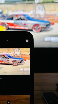 A colorful race car is displayed on a screen and simultaneously captured on a smartphone. The car features vibrant red and blue colors with decals. It is parked in what seems to be a race track environment with barriers and indistinct background structures.