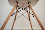 A close-up view of the underside of a chair, focusing on the wooden legs and metal supports. The legs are light-colored wood with a smooth finish, connected by black metal rods forming a geometric pattern.