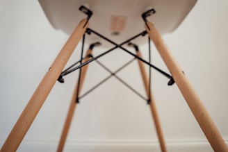 A close-up view of the underside of a chair, focusing on the wooden legs and metal supports. The legs are light-colored wood with a smooth finish, connected by black metal rods forming a geometric pattern.