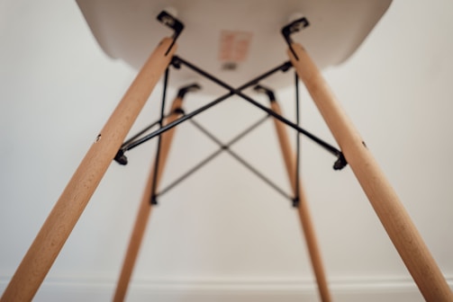 A close-up view of the underside of a chair, focusing on the wooden legs and metal supports. The legs are light-colored wood with a smooth finish, connected by black metal rods forming a geometric pattern.