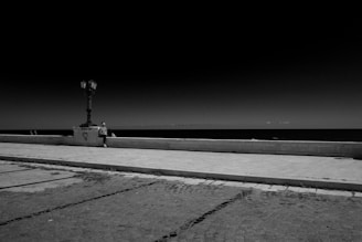 A solitary person sits on a stone bench beside a lamppost along a desolate waterfront promenade. The vast, dark sky looms over an equally dark sea in the background, while the foreground features a textured stone paved walkway. A heart graffiti is visible on the lamppost's base.