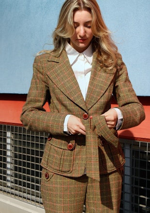 A person with long, wavy hair is wearing a tailored plaid suit with a double-breasted jacket and matching trousers. The suit has red-brown buttons and the person is adjusting the jacket. The background consists of a blue wall above a red strip with a metal grid.