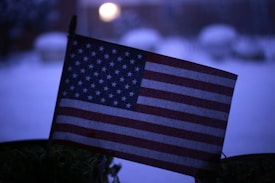 A small American flag with stars and stripes is prominently displayed against a blurred background. The scene appears to be set during winter, as there are hints of snow visible and the overall tone is cool and dark, possibly taken at dusk or dawn.