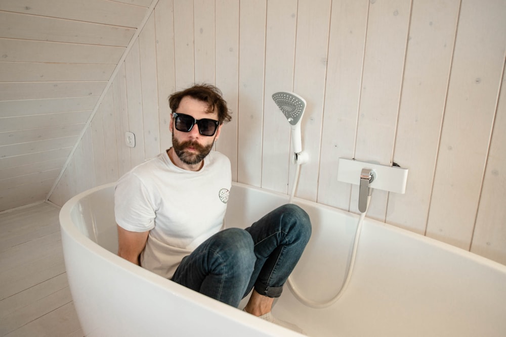 man in white crew neck t-shirt and blue denim jeans sitting on white ceramic bathtub