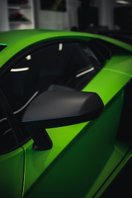 Close-up view of a sleek and modern side mirror of a bright green sports car. The car's sharp lines and glossy finish are evident, with the mirror featuring a carbon fiber texture. In the background, blurry interiors of a showroom can be seen.