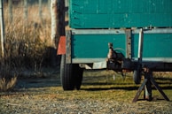 A green wooden trailer is parked on a grassy and gravel surface, with a metal hitch and support structures visible. The background includes tall grass and a fence, suggesting a rural or outdoor setting.