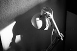 A black and white photograph featuring a desk lamp casting a shadow on a textured wall. The lamp is positioned at an angle, resulting in a distinct play of light and shadow.