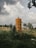 An old, rusted cylindrical tank stands in a grassy field with tall plants growing around it. The sky above is filled with large, fluffy clouds, and there are trees and a fence in the background. A small structure can be seen in the distance.