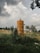An old, rusted cylindrical tank stands in a grassy field with tall plants growing around it. The sky above is filled with large, fluffy clouds, and there are trees and a fence in the background. A small structure can be seen in the distance.