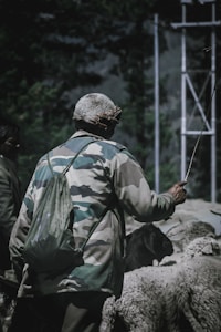 A person wearing a camouflage jacket and a backpack holds a stick while standing among a herd of sheep. The setting appears rural, with metal structures visible in the background against a backdrop of foliage.