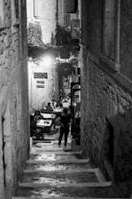 A narrow, dimly lit alleyway with stone steps leading down to an open-air dining area. People are seated at tables enjoying a meal, with the scene giving a sense of intimacy. The walls are made of stone, and foliage can be seen at the top of the alley. Signs with Italian words are visible, enhancing an old-world charm.