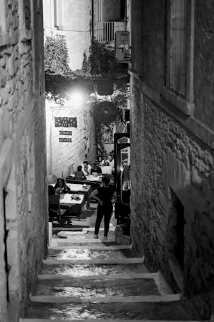 A narrow, dimly lit alleyway with stone steps leading down to an open-air dining area. People are seated at tables enjoying a meal, with the scene giving a sense of intimacy. The walls are made of stone, and foliage can be seen at the top of the alley. Signs with Italian words are visible, enhancing an old-world charm.