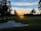 A serene landscape featuring a golf course at sunset, with the sun casting a warm glow across the sky. Tall trees line the fairway, their silhouettes dark against the colorful horizon. A sand trap is visible in the foreground, blending into the lush green grass. The sky is a mix of blues and oranges as the day transitions to night.