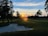 A serene landscape featuring a golf course at sunset, with the sun casting a warm glow across the sky. Tall trees line the fairway, their silhouettes dark against the colorful horizon. A sand trap is visible in the foreground, blending into the lush green grass. The sky is a mix of blues and oranges as the day transitions to night.
