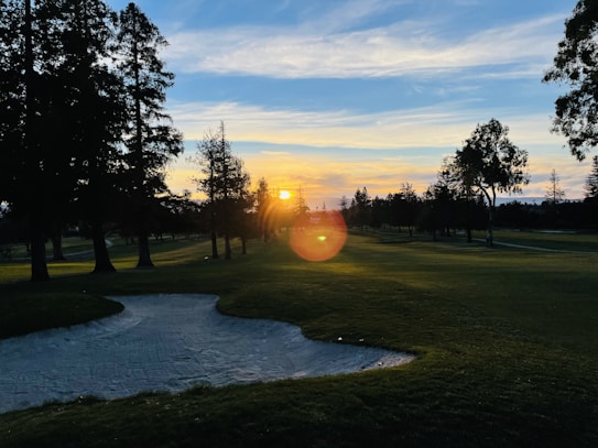 A serene landscape featuring a golf course at sunset, with the sun casting a warm glow across the sky. Tall trees line the fairway, their silhouettes dark against the colorful horizon. A sand trap is visible in the foreground, blending into the lush green grass. The sky is a mix of blues and oranges as the day transitions to night.
