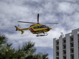 A helicopter with a yellow and red color scheme is flying near a tall building with multiple windows. The helicopter is a model used for civil security. The sky is filled with clouds, and there are green branches in the bottom left corner.