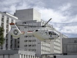 A medical helicopter is in mid-flight near a large, modern hospital building. The helicopter is white with red accents and displays the word 'SAMU' on its side, indicating it is an emergency medical service vehicle. The background consists of the hospital's geometric, rectangular architecture under a cloudy sky.