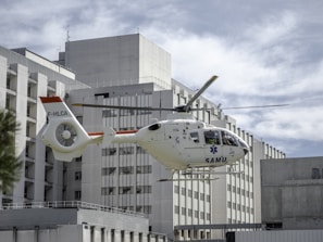 A medical helicopter is in mid-flight near a large, modern hospital building. The helicopter is white with red accents and displays the word 'SAMU' on its side, indicating it is an emergency medical service vehicle. The background consists of the hospital's geometric, rectangular architecture under a cloudy sky.